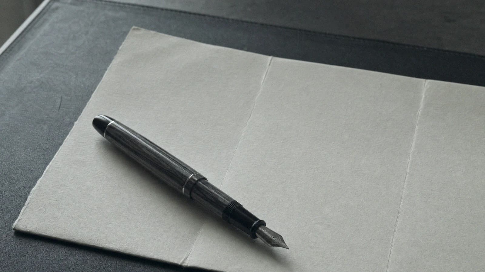 Pióro wieczne na złożonym arkuszu papieru, na ciemnym blacie w chłodnym świetle z okna